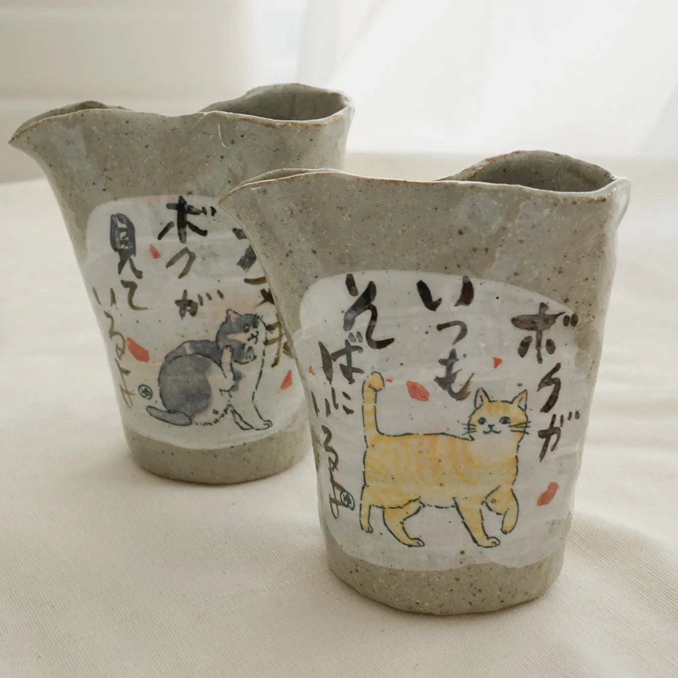 Two handmade ceramic cat-shaped cups with a speckled gray-beige glaze, featuring white labels with Japanese text and illustrations of cats in black, gray, and yellow, set against a soft white background.