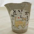 A handmade ceramic pitcher with a wavy rim, featuring a speckled gray glaze and hand-painted black calligraphy with a small yellow accent on the front, set against a light fabric background.