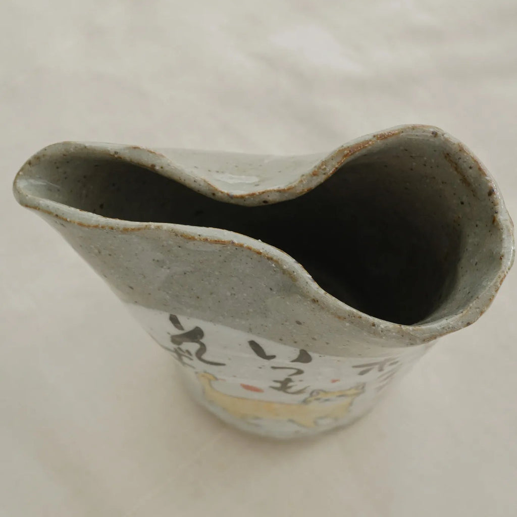 A handmade ceramic pitcher with a wavy rim, featuring a speckled gray glaze and hand-painted black calligraphy with a small yellow accent on the front, set against a light fabric background.