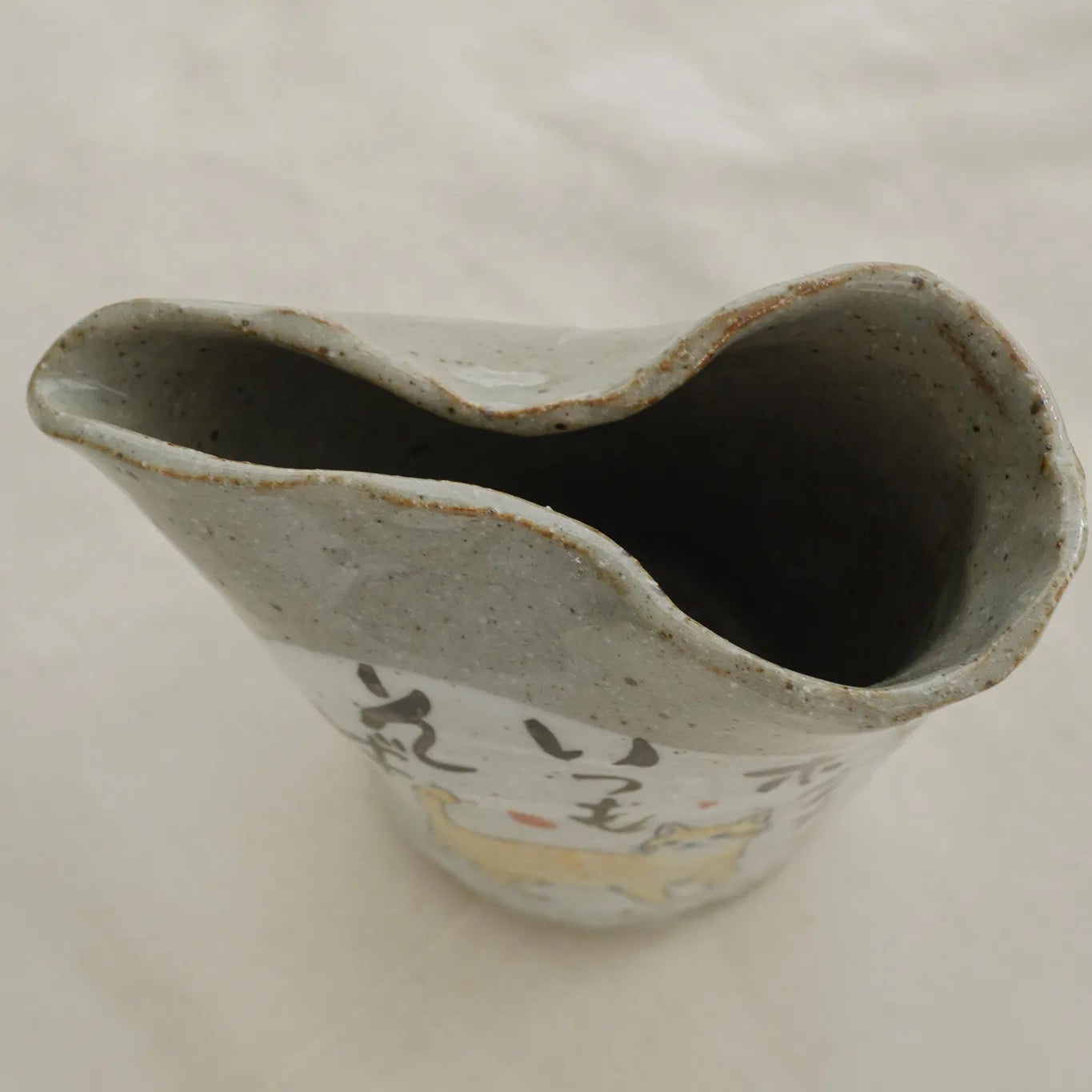 A handmade ceramic pitcher with a wavy rim, featuring a speckled gray glaze and hand-painted black calligraphy with a small yellow accent on the front, set against a light fabric background.