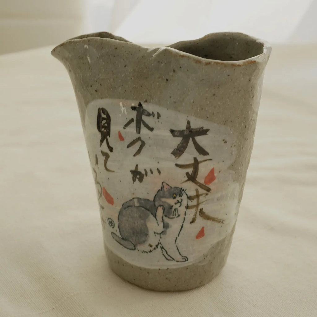 A handmade Japanese ceramic pitcher with a textured gray-beige glaze, featuring a hand-painted cat illustration and Japanese calligraphy on a white label.
