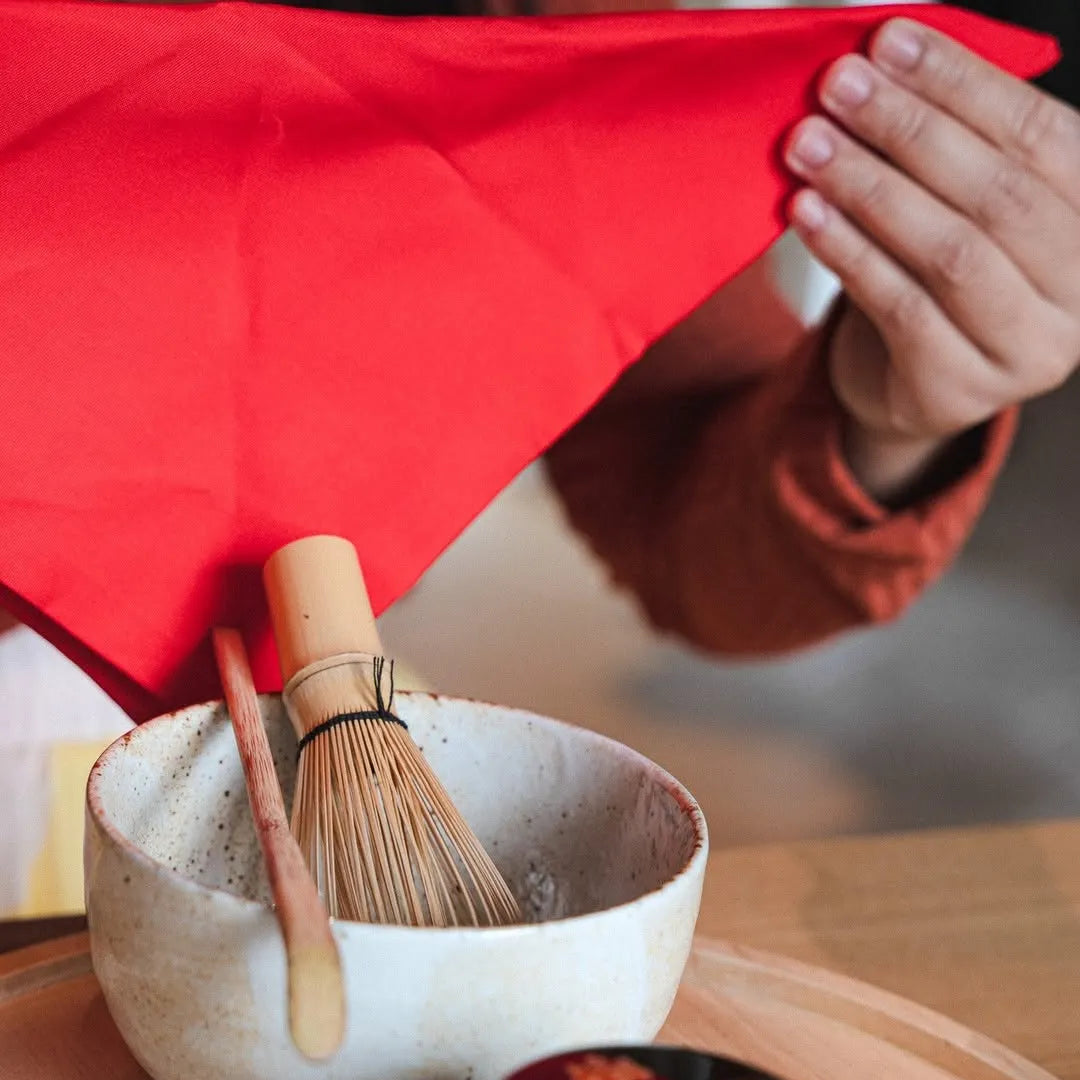 Ceramic bowl with wooden whisk and black container on a wooden surface, person holding red cloth in background